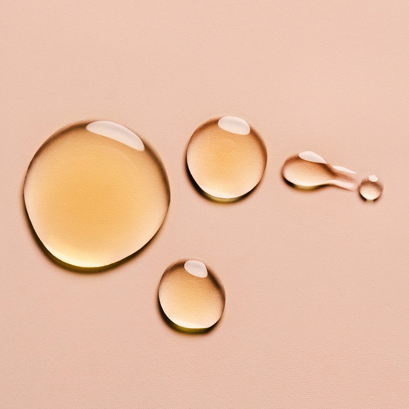 Close-up shot of golden liquid drops with a glossy finish on a neutral beige background, suitable for beauty or product imagery.