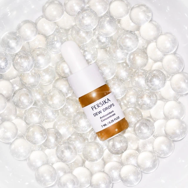 Persika Dew Drops antioxidant concentrate bottle with dropper on white marble balls. Beauty product photography for skincare.