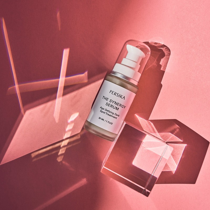 Persika The Synergy Serum bottle with pump, clear cap, and box on a red background with light reflections, skincare product.