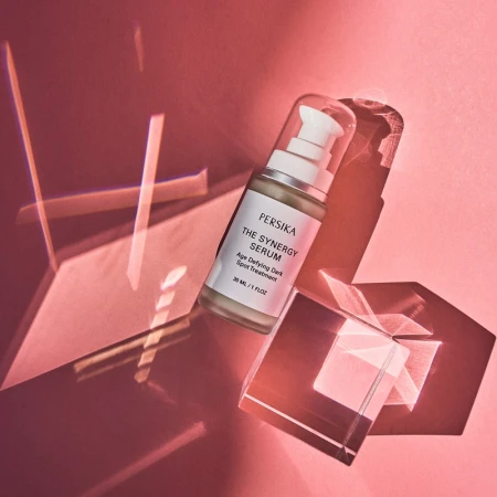 Persika The Synergy Serum bottle with pump, clear cap, and box on a red background with light reflections, skincare product.