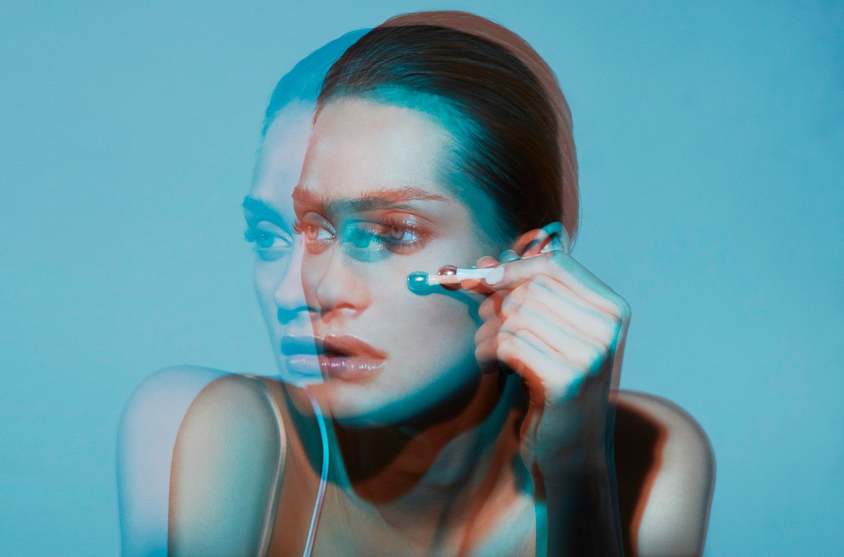 Close-up of a woman applying makeup with a blue and red duotone effect against a blue background in a beauty style photograph.