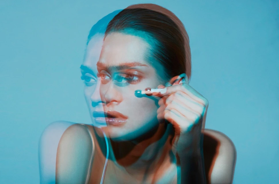 Close-up of a woman applying makeup with a blue and red duotone effect against a blue background in a beauty style photograph.