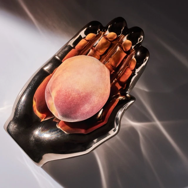 Close-up studio shot of a fresh peach resting in a decorative metallic hand dish, catching light and shadows on a light background.