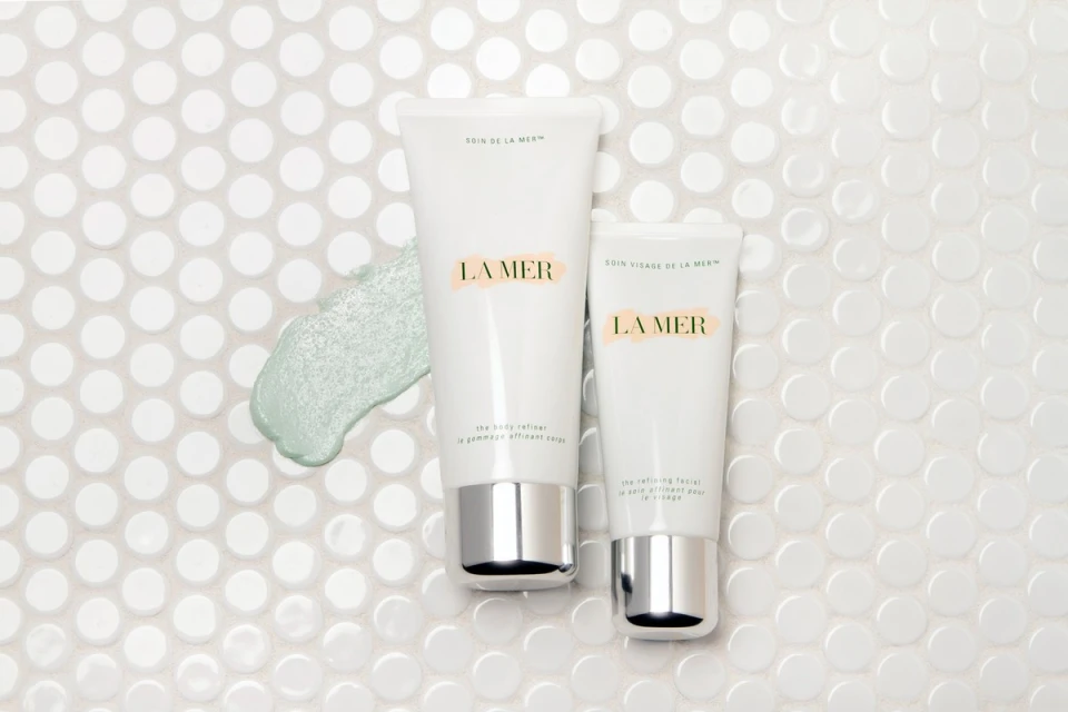Close-up of La Mer skincare products against a bright white mosaic tile background, highlighting the brand's body and face refiner creams.