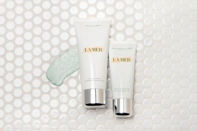 Close-up of La Mer skincare products against a bright white mosaic tile background, highlighting the brand's body and face refiner creams.
