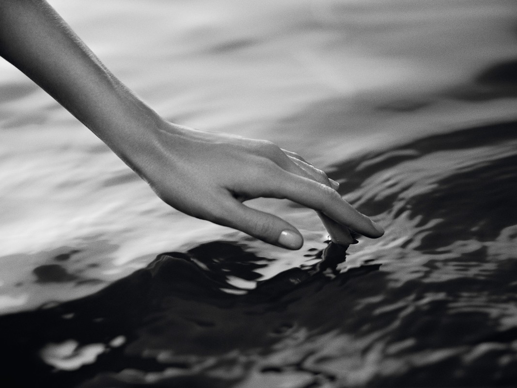 Close-up of a human arm and hand gently touching the rippled surface of water in a black and white artistic style.