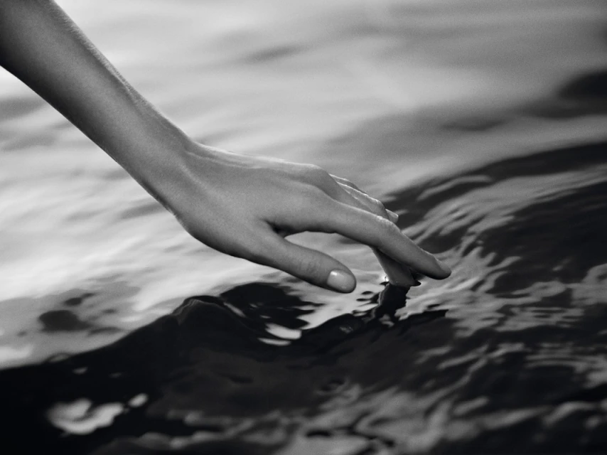 Close-up of a human arm and hand gently touching the rippled surface of water in a black and white artistic style.