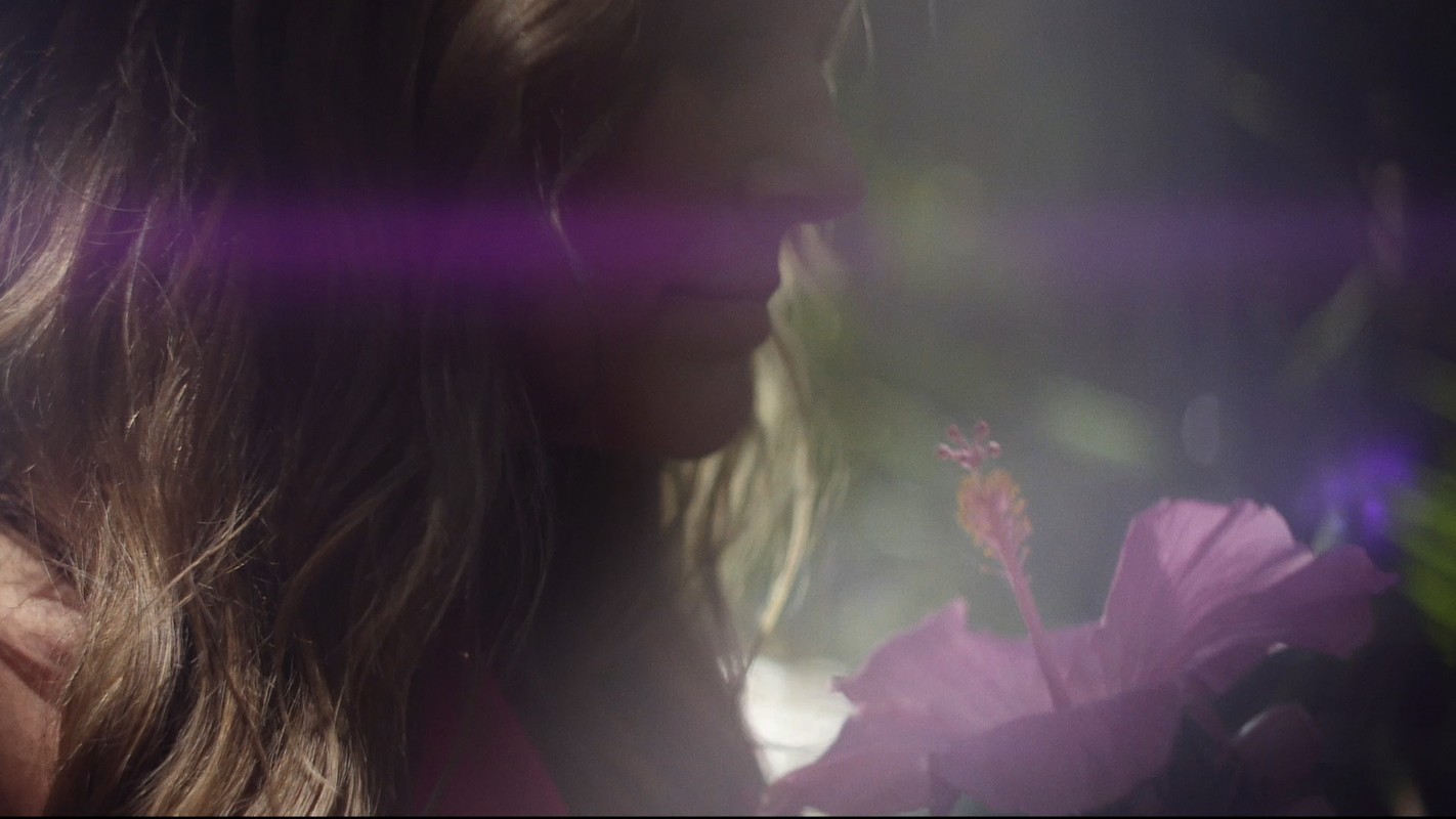 Woman with long blonde hair in soft focus, standing outdoors, next to a pink hibiscus flower with green foliage in the background.