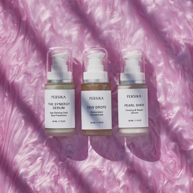 Three Persika serum bottles lined up against a pink swirling background. The product names include Dew Drops, Pearl Shen, and Synergy Serum.