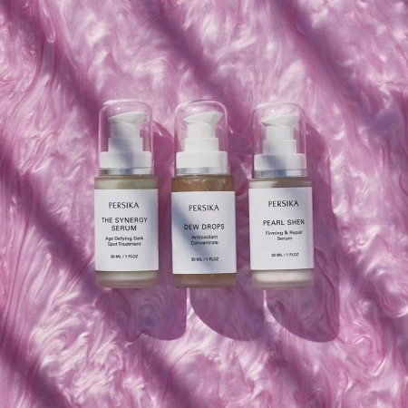 Three Persika serum bottles lined up against a pink swirling background. The product names include Dew Drops, Pearl Shen, and Synergy Serum.