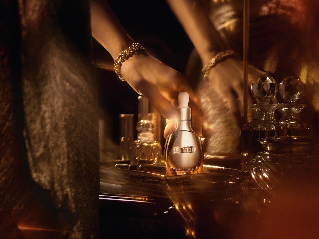 Glimmering close-up of La Mer serum bottle on a reflective surface with a woman's hand and crystal perfume bottles.