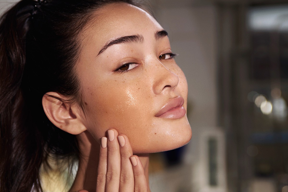 Close-up portrait of an Asian woman applying moisturizer to her face, highlighting healthy skincare and a radiant complexion in soft light.