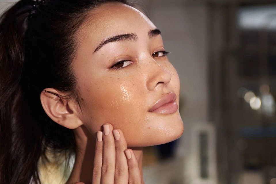 Close-up portrait of an Asian woman applying moisturizer to her face, highlighting healthy skincare and a radiant complexion in soft light.