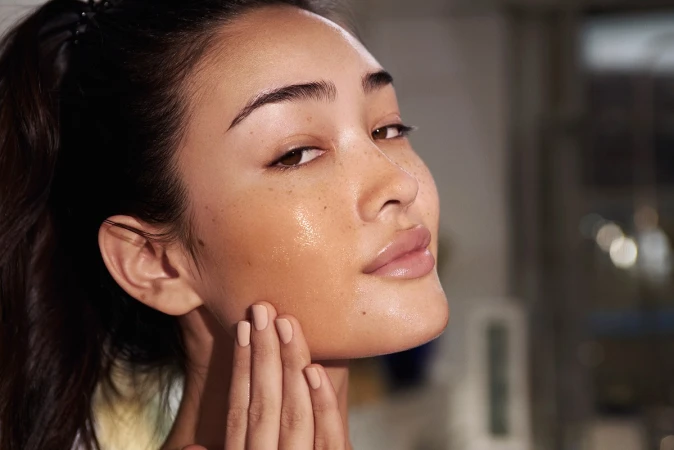 Close-up portrait of an Asian woman applying moisturizer to her face, highlighting healthy skincare and a radiant complexion in soft light.