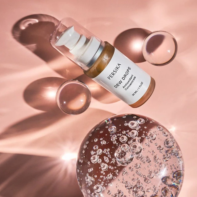 Persika Dew Drops antioxidant concentrate cosmetic serum bottle displayed with transparent spheres against a light pink backdrop.
