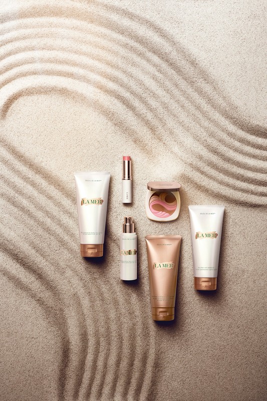 La Mer skincare and makeup products arranged on sand featuring bronzer, lipstick, serum, cleanser, and cream in a high-angle shot.