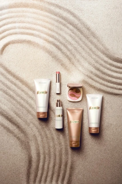 La Mer skincare and makeup products arranged on sand featuring bronzer, lipstick, serum, cleanser, and cream in a high-angle shot.