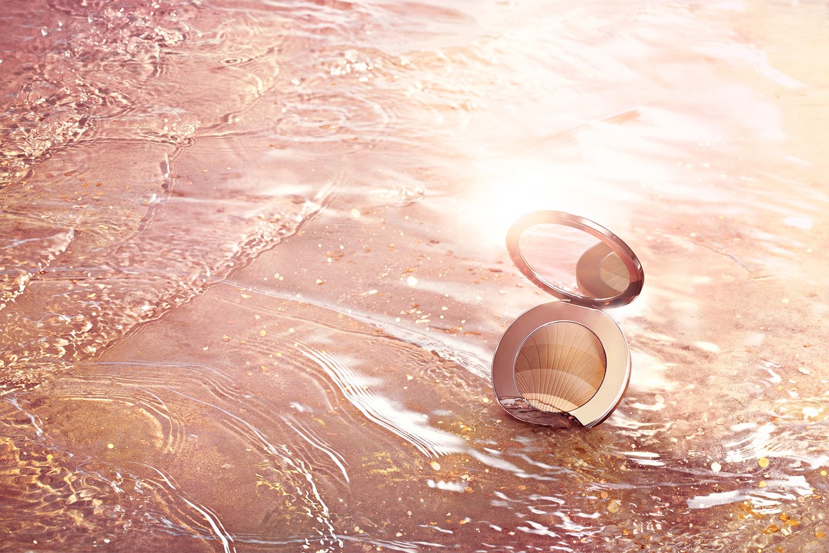 Makeup powder compact sits open on a sunlit beach with water gently lapping over the sand, creating a soft, cosmetic product shot.