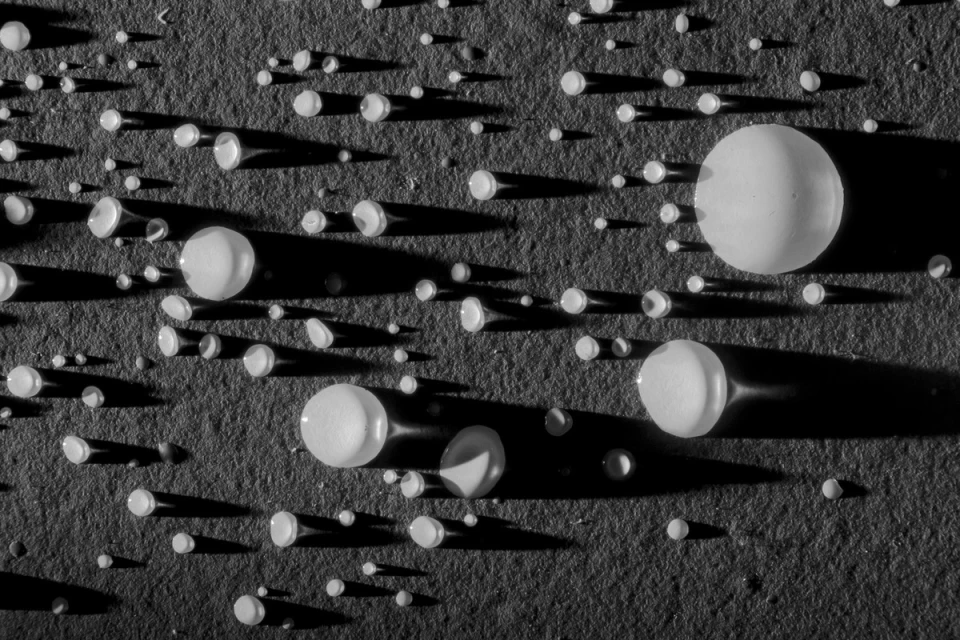 High-angle close-up shot of water droplets on gray textured surface with long shadows, creating a textured, abstract, monochromatic background.