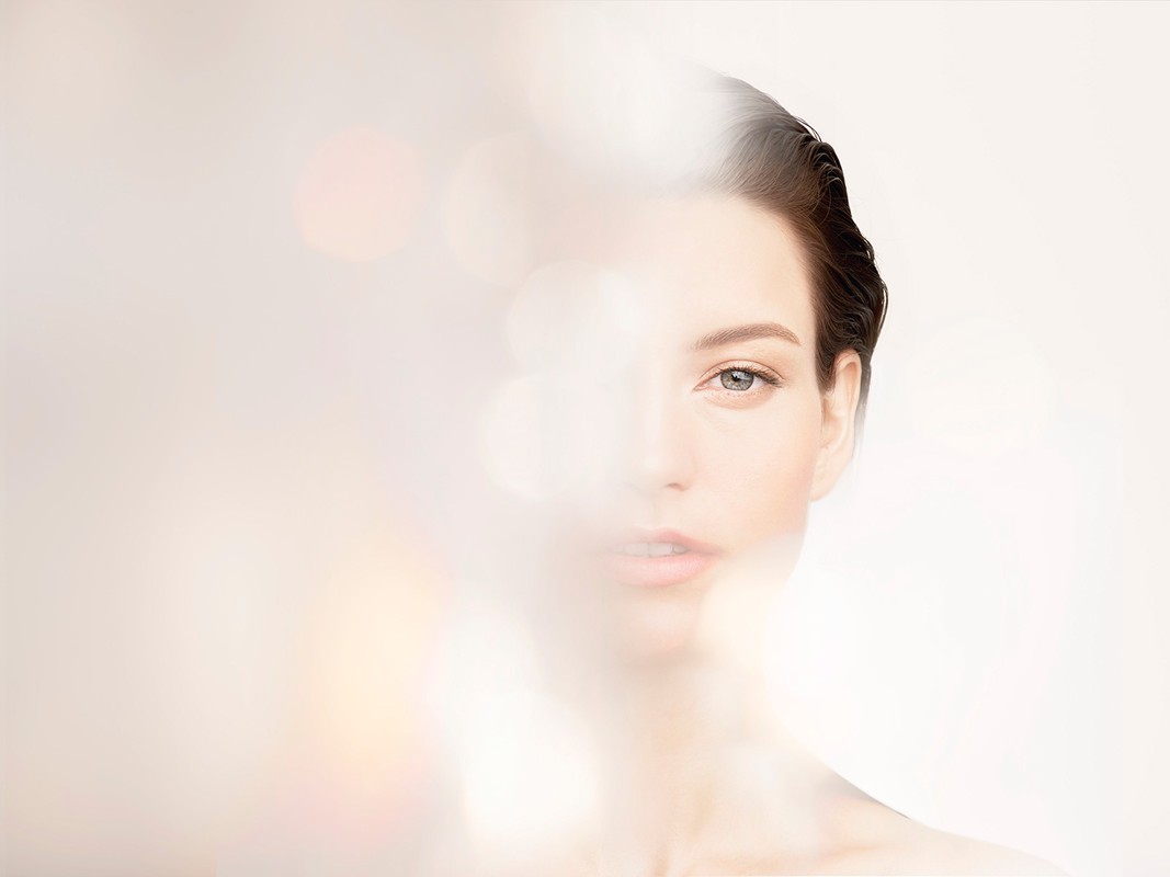 Close-up portrait of a serene adult woman with smooth skin, softly lit with a blurred bokeh effect in the foreground and neutral background.