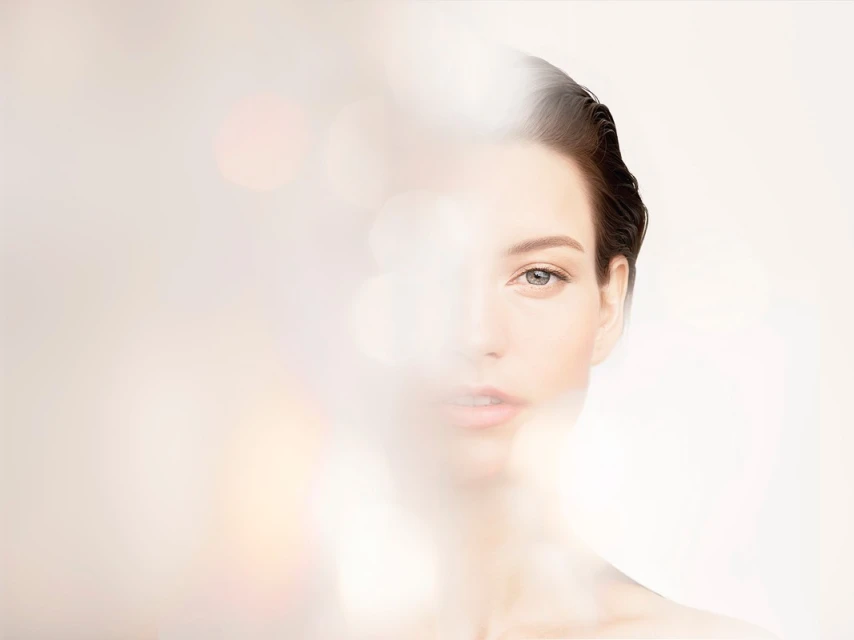 Close-up portrait of a serene adult woman with smooth skin, softly lit with a blurred bokeh effect in the foreground and neutral background.