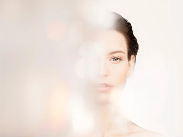 Close-up portrait of a serene adult woman with smooth skin, softly lit with a blurred bokeh effect in the foreground and neutral background.