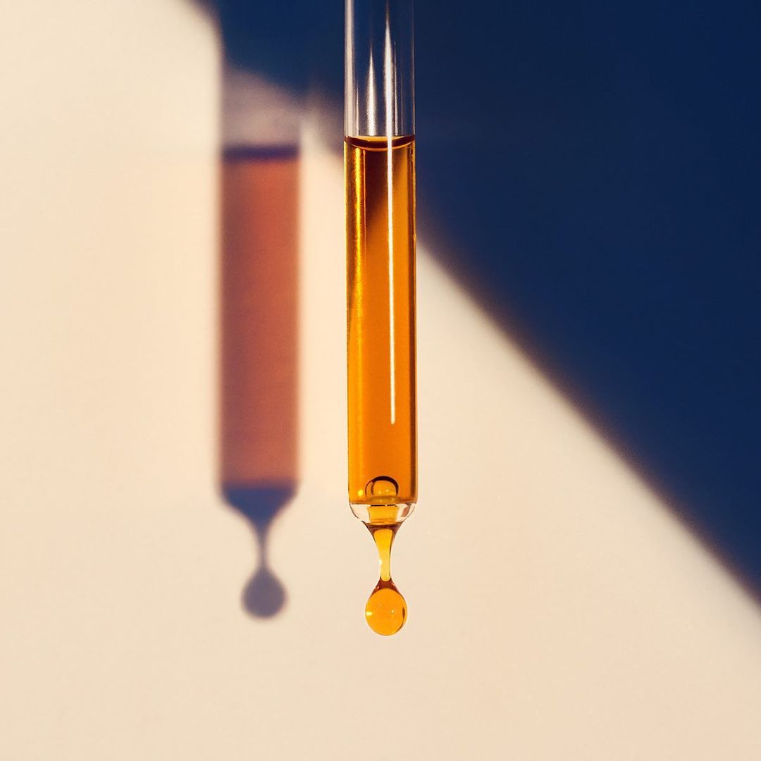 Close-up studio shot of a clear pipette dispensing a single golden liquid drop with a geometric shadow on a neutral background.