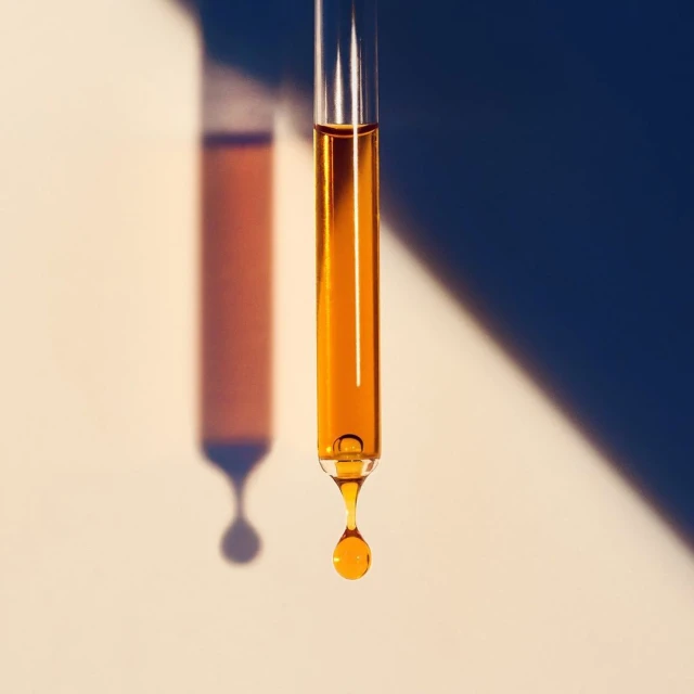 Close-up studio shot of a clear pipette dispensing a single golden liquid drop with a geometric shadow on a neutral background.