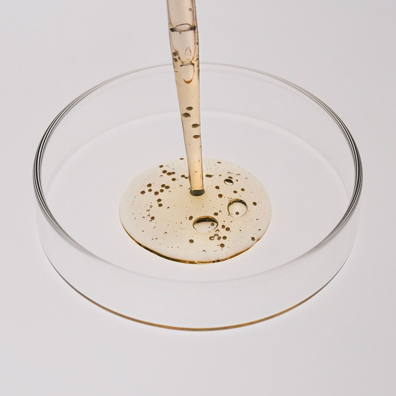 Close-up of a pipette dispensing golden serum into a glass petri dish against a white background, showcasing beauty and healthcare concepts.