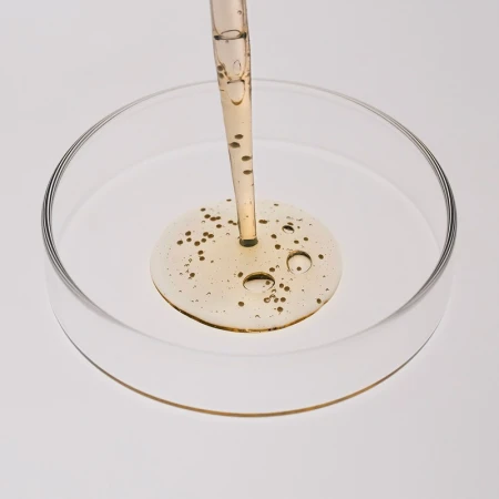 Close-up of a pipette dispensing golden serum into a glass petri dish against a white background, showcasing beauty and healthcare concepts.