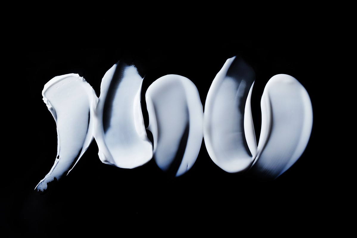 Close-up of thick, pale, blue-tinged cream streaks on a solid black background, resembling cosmetic smears or abstract brushstrokes.