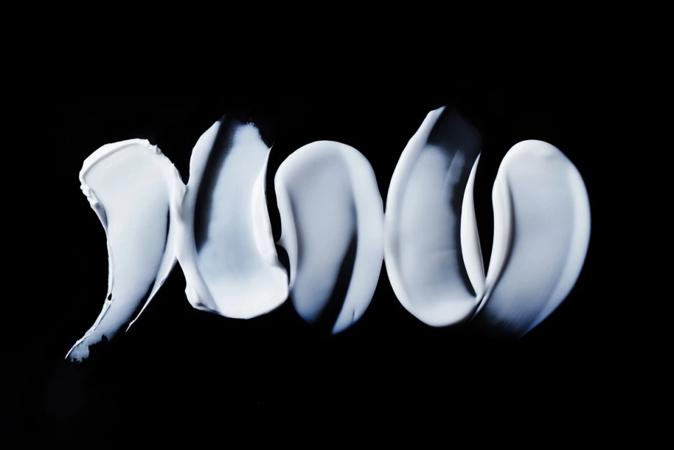 Close-up of thick, pale, blue-tinged cream streaks on a solid black background, resembling cosmetic smears or abstract brushstrokes.