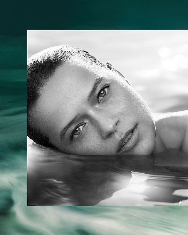Monochrome close-up of an adult female partially submerged in water, eyes open, with an abstract wave pattern in the background.