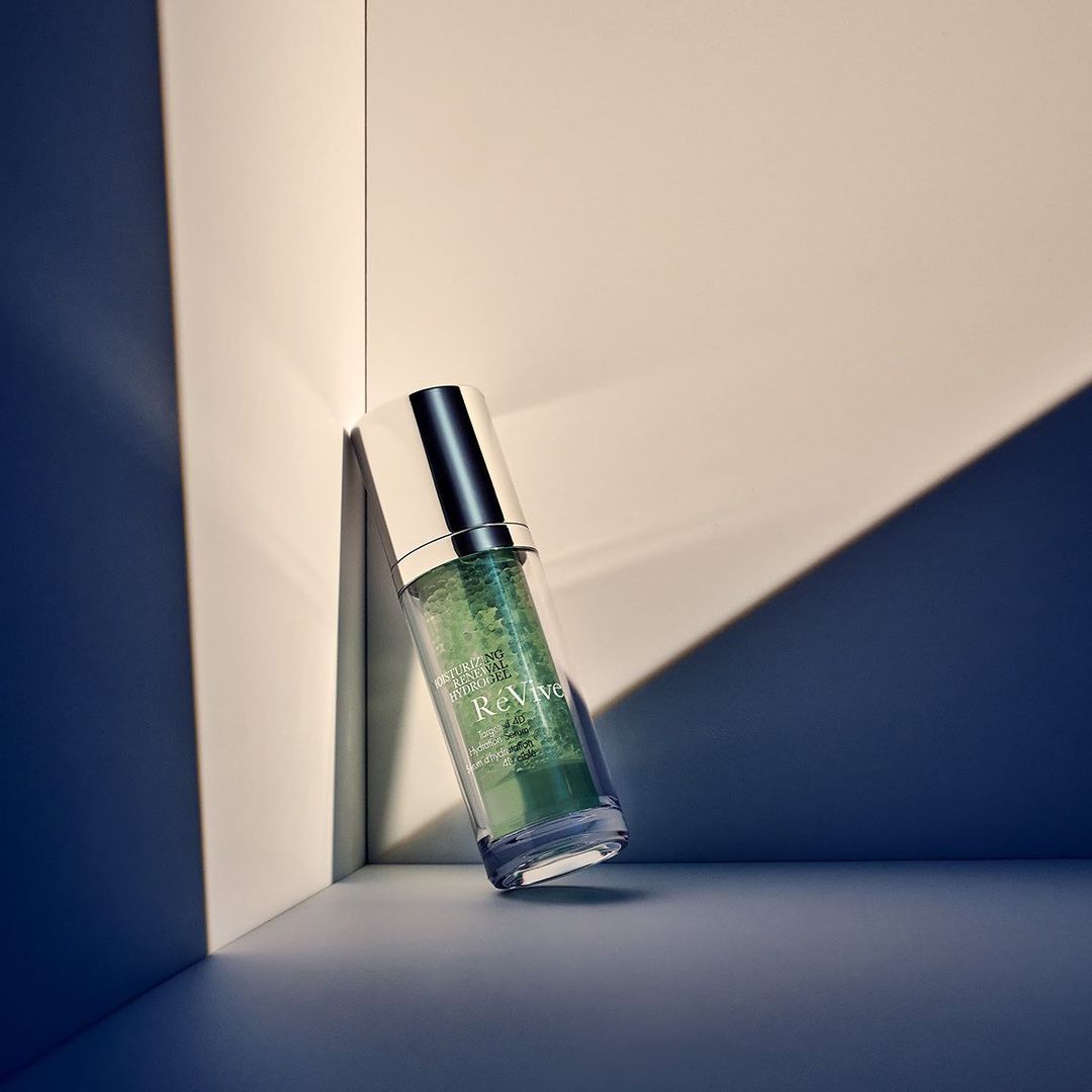 Serum bottle by white wall. Clear bottle with silver top filled with green liquid, sits against white wall, shadows cast on backdrop.