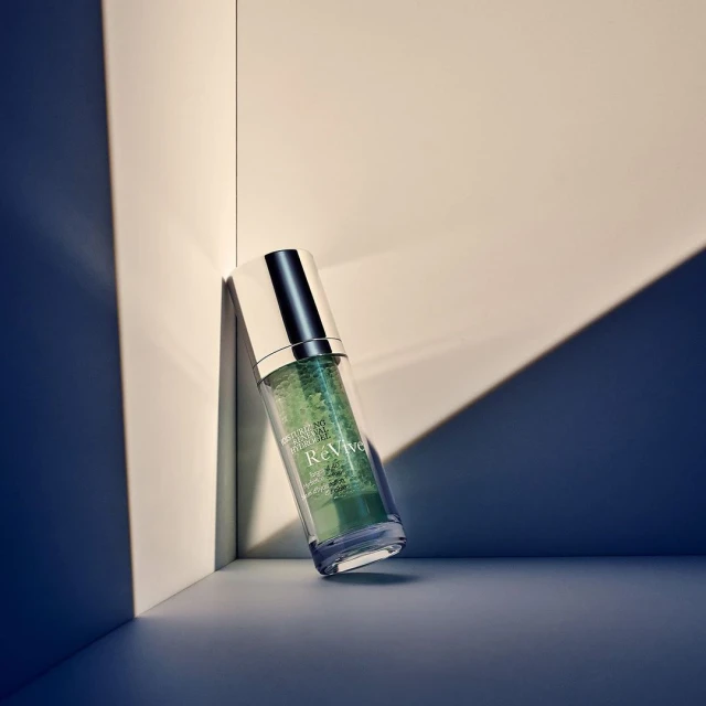Serum bottle by white wall. Clear bottle with silver top filled with green liquid, sits against white wall, shadows cast on backdrop.