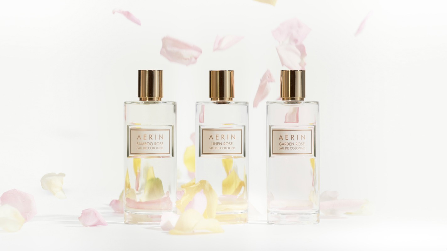 Three Aerin perfume bottles centered on a white background with pale pink and yellow petals gently falling around them. Minimalist design.