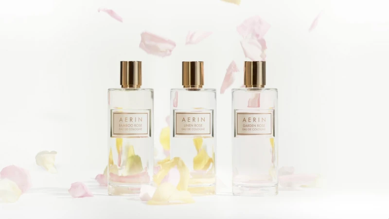 Three Aerin perfume bottles centered on a white background with pale pink and yellow petals gently falling around them. Minimalist design.