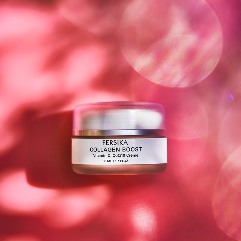 Product shot of Persika Collagen Boost cream jar with a transparent lid on a pink background with blurred light patterns.