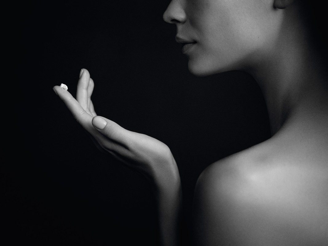 Close-up black and white studio shot of a fair-skinned woman's hand holding a small white cream against a dark background.
