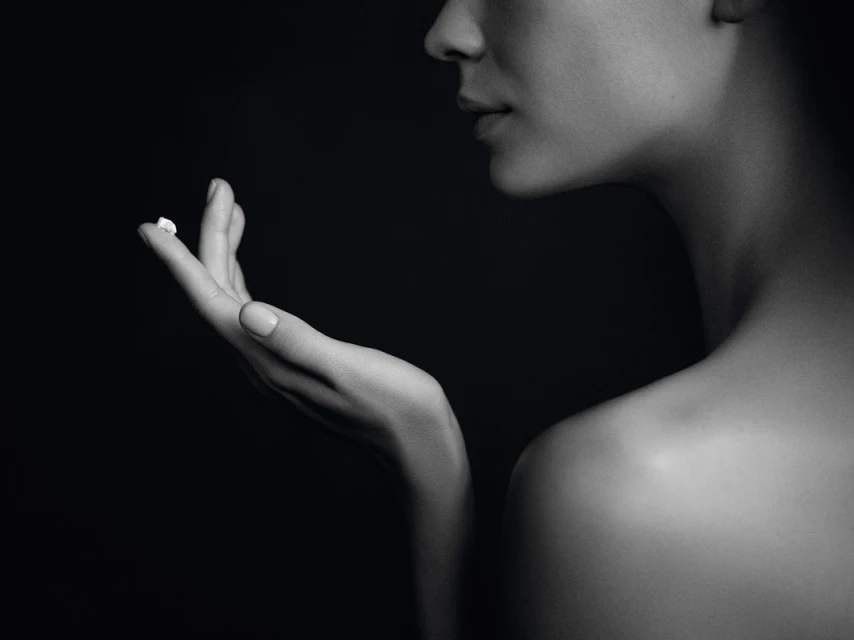 Close-up black and white studio shot of a fair-skinned woman's hand holding a small white cream against a dark background.