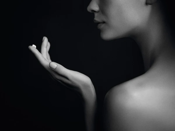 Close-up black and white studio shot of a fair-skinned woman's hand holding a small white cream against a dark background.
