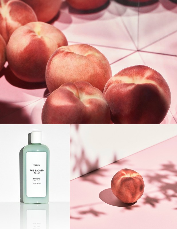 Still life of ripe peaches, arranged alongside a turquoise bottle, set on a pink surface under a diffused light source creating soft shadows.