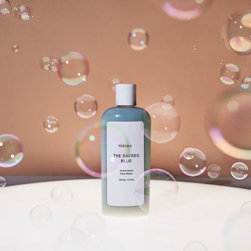 Blue Persika face wash bottle sits on a white surface surrounded by bubbles against a peach background in a beauty product shot.