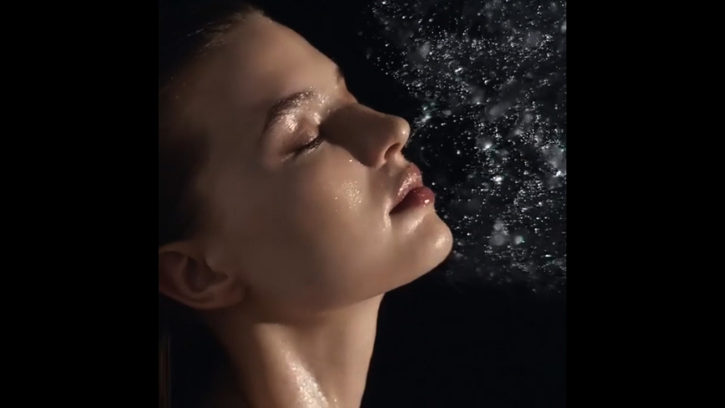 Close-up shot of a young woman with wet skin, posed in front of a dark background with splashing water droplets, focusing on beauty.