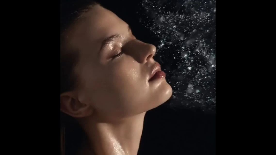 Close-up shot of a young woman with wet skin, posed in front of a dark background with splashing water droplets, focusing on beauty.