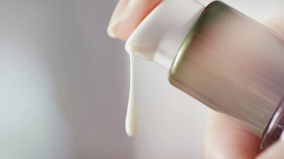 Close-up of a person's hand dispensing white cosmetic cream from a sleek, modern bottle with green and gray accents against a neutral backdrop.