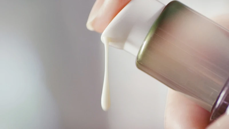 Close-up of a person's hand dispensing white cosmetic cream from a sleek, modern bottle with green and gray accents against a neutral backdrop.