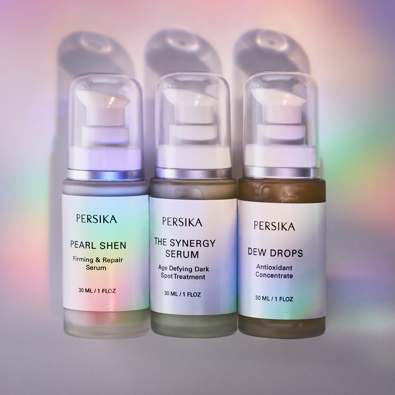 Three Persika serum bottles lined up against a soft, rainbow-hued gradient background. Each bottle features a pump dispenser and a label.