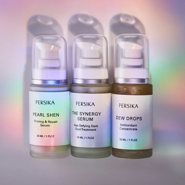 Three Persika serum bottles lined up against a soft, rainbow-hued gradient background. Each bottle features a pump dispenser and a label.