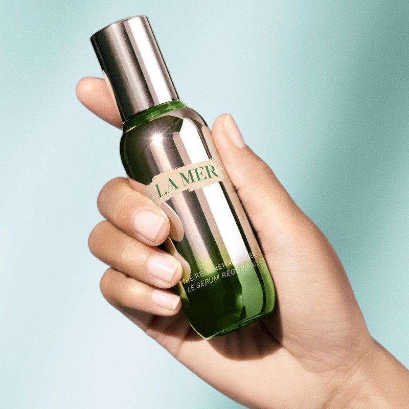 Hand holds La Mer serum bottle, showcasing skincare product against a soft teal background in a beauty or fashion context.